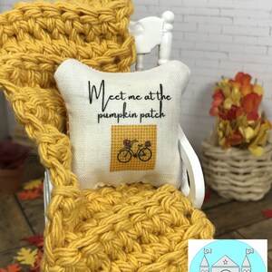 Op de afbeelding: Een wit kussen met de tekst "Meet me at the pumpkin patch" en een klein beeld van een fiets met een mand vol bloemen. Het kussen ligt op een geel gehaakt deken op een witte stoel.