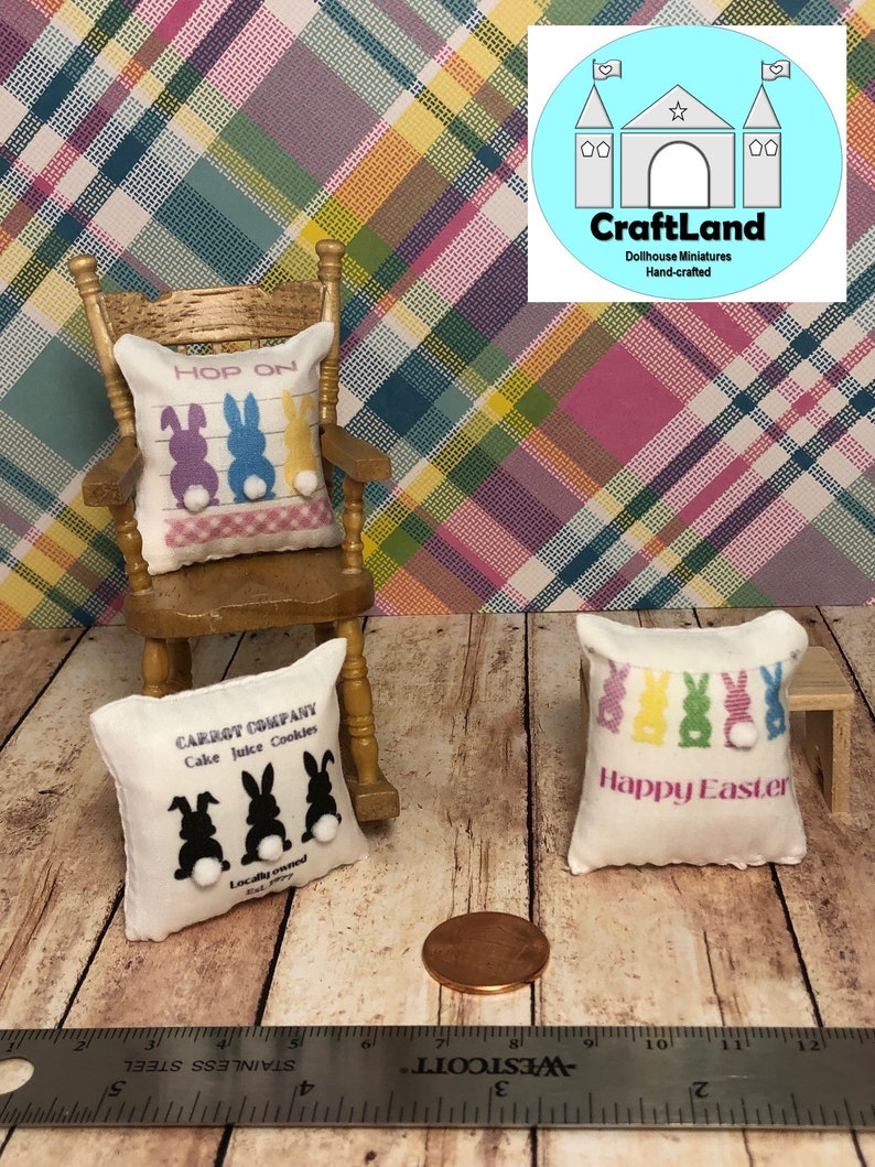 May include: Three miniature pillows with Easter themes. The first pillow is white with black bunny silhouettes and the text "CARROT COMPANY Cake Juice Cookies Locally owned". The second pillow is white with a pink, blue, and yellow bunny silhouette and the text "HOP ON". The third pillow is white with a multi-colored bunny silhouette and the text "Happy Easter".