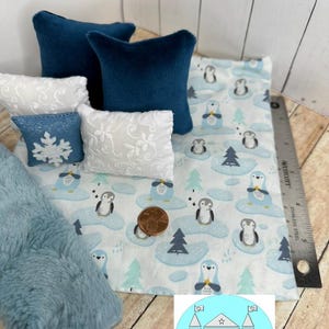 Op de afbeelding: Een miniatuur poppenhuisbed met beddengoed met een blauw en wit pingu&iuml;npatroon. Het bed heeft twee blauwe kussens en twee witte kussens met een kantpatroon. Er ligt een blauwe nep bontdeken op het bed.