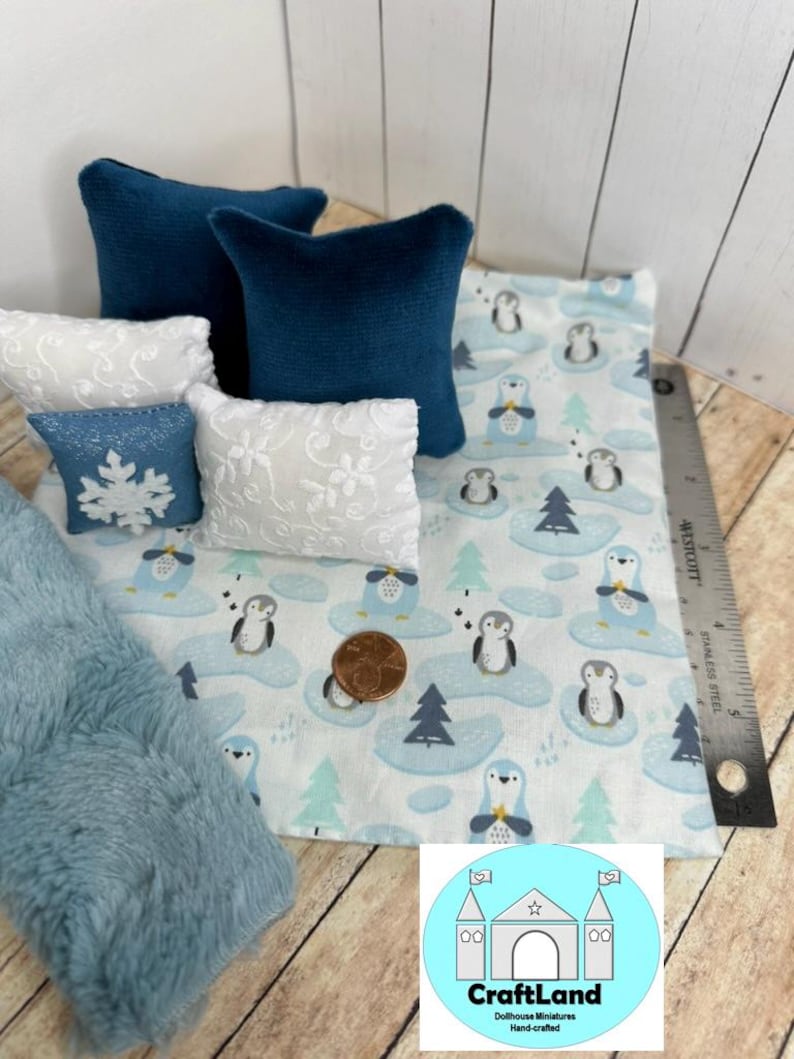 Op de afbeelding: Een miniatuur poppenhuisbed met beddengoed met een blauw en wit pingu&iuml;npatroon. Het bed heeft twee blauwe kussens en twee witte kussens met een kantpatroon. Er ligt een blauwe nep bontdeken op het bed.