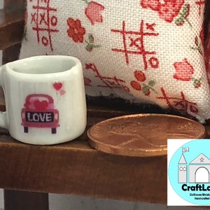 May include: White ceramic miniature coffee mug with a pink car design and the word "LOVE" on the back. The mug is sitting on a wooden surface next to a penny.