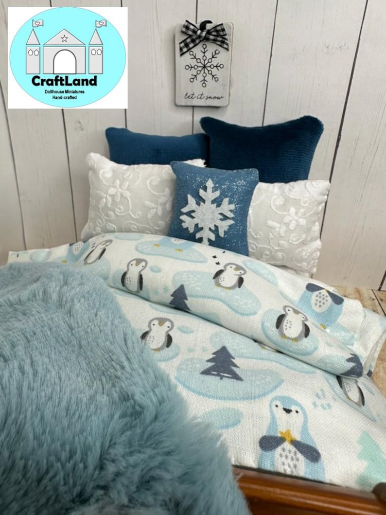 Op de afbeelding: Een poppenbed met een blauwe en witte beddengoedset met pingu&iuml;nmotief. Het bed heeft een blauwe nep bontdeken en verschillende witte en blauwe kussens. Een bord boven het bed staat "Let it snow" met een sneeuwvlokkenontwerp.