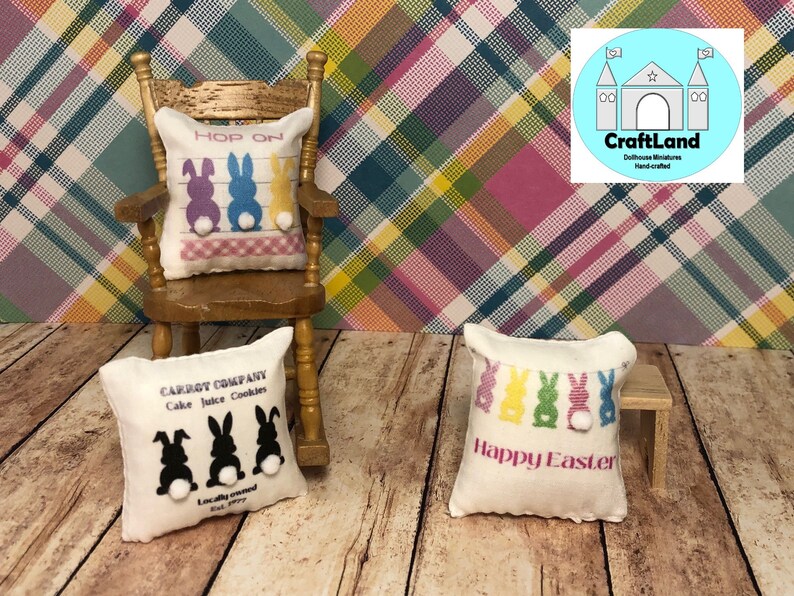 May include: Three miniature pillows with Easter bunny designs. The first pillow is white with black bunnies and the text "CARROT COMPANY Cake Juice Cookies Locally owned Est. Perry". The second pillow is white with colorful bunnies and the text "HOP ON". The third pillow is white with colorful bunnies and the text "Happy Easter".