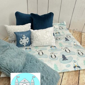 Op de afbeelding: Een blauwe en witte deken met een pingu&iuml;npatroon, een blauwe nep bontdeken en vier kussens. Twee van de kussens zijn diep tealblauw, &eacute;&eacute;n is wit met een sneeuwvlokkenpatroon en &eacute;&eacute;n is wit met een bloemenpatroon.