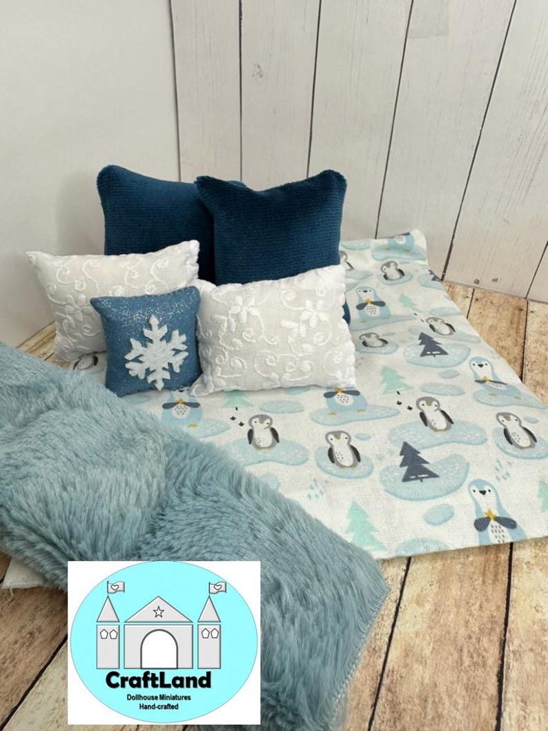 Op de afbeelding: Een blauwe en witte deken met een pingu&iuml;npatroon, een blauwe nep bontdeken en vier kussens. Twee van de kussens zijn diep tealblauw, &eacute;&eacute;n is wit met een sneeuwvlokkenpatroon en &eacute;&eacute;n is wit met een bloemenpatroon.