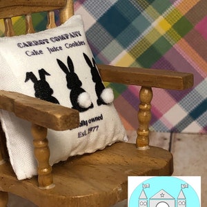 May include: A white pillow with black bunny silhouettes and the text "CARROT COMPANY Cake Juice Cookies" and "ally owned Est. 1977". The pillow is sitting on a wooden rocking chair with a light blue background.