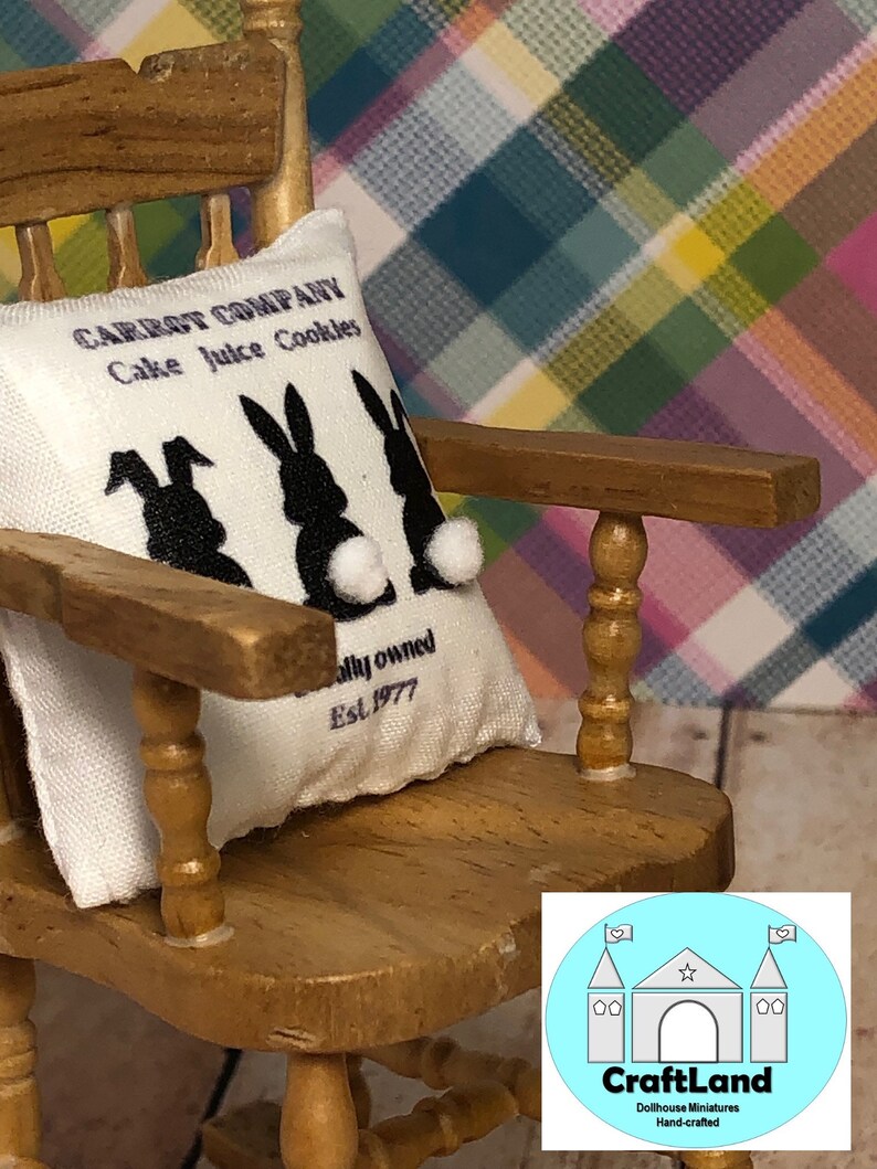 May include: A white pillow with black bunny silhouettes and the text "CARROT COMPANY Cake Juice Cookies" and "ally owned Est. 1977". The pillow is sitting on a wooden rocking chair with a light blue background.