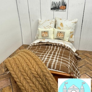 May include: A miniature dollhouse bed with a brown and white plaid comforter, a brown knitted blanket, and two white pillows with embroidered pumpkins. The pillows have a white background with brown wheat stalks.