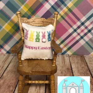 May include: A miniature rocking chair with a white pillow featuring colorful Easter bunnies and the text "Happy Easter". The chair is on a wooden floor with a plaid background.
