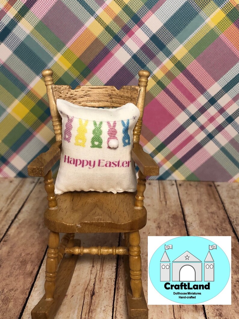 May include: A miniature rocking chair with a white pillow featuring colorful Easter bunnies and the text "Happy Easter". The chair is on a wooden floor with a plaid background.