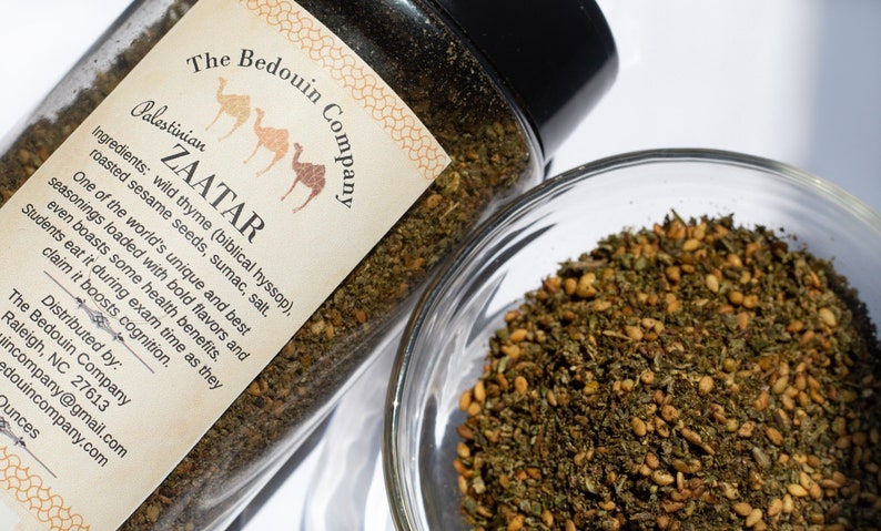 Może przedstawiać: Szklany słoik wypełniony mieszanką zi&oacute;ł i przypraw z etykietą "The Bedouin Company Palestinian ZAATAR". Etykieta zawiera r&oacute;wnież listę składnik&oacute;w: dziki tymianek, nasiona sezamu, sumak i s&oacute;l. Słoik stoi na białym tle.