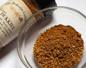 May include: A clear glass bowl filled with dukkah, a Middle Eastern spice blend. A jar of The Bedouin Company dukkah is partially visible, with text listing ingredients: thyme, sumac, sesame seeds, chickpeas, cumin, and salt.