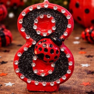 May include: A red number eight candle with a ladybug design. The candle is decorated with black glitter and clear rhinestones. A ladybug is attached to the front of the candle. The candle is on a wooden surface with star-shaped confetti.