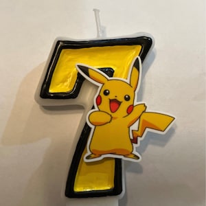 May include: A number 7 birthday candle with a yellow Pikachu graphic. The candle is yellow with a black outline and a white base. The Pikachu graphic is yellow, red, and black.
