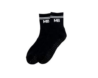 May include: A pair of black socks with white stripes at the top and the letters "MB" in white. The soles have small, textured grips. These socks are suitable for sports or everyday use.