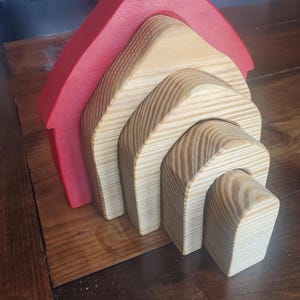 May include: A wooden stacking toy shaped like a house with a red roof. The house is made of five pieces that fit inside each other.