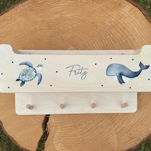 Könnte beinhalten: Wandregal aus hellem Holz mit einer Schildkröten- und Walillustration. Der Name "Fritz" ist kursiv geschrieben. Enthält drei Holzstifte zum Aufhängen von Gegenständen. Ein dekoratives Stück für ein Kinderzimmer.