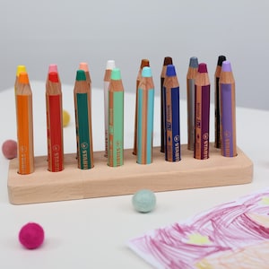 May include: A set of colorful pencils in a wooden holder. The pencils are various colors, including orange, red, green, and blue. Small felt balls in shades of pink and blue are scattered around the pencils. A child's drawing is visible in the corner.