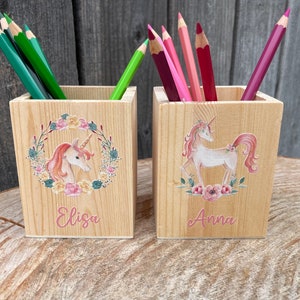 Puede incluir: Dos porta lápices de madera con diseños de unicornio. El porta lápices de la izquierda tiene una cabeza de unicornio con una corona de flores y el nombre "Eliza" impreso debajo. El porta lápices de la derecha tiene un unicornio completo con un diseño floral y el nombre "Anna" impreso debajo.
