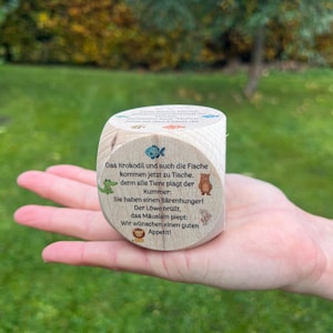 Può includere: Un blocco di legno con bordi arrotondati, con testo stampato e piccole illustrazioni di animali. Il testo è in tedesco e include un pesce, un coccodrillo, un orso e un topo. Il blocco è tenuto in mano su uno sfondo verde sfocato.