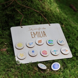 Peut inclure: Un panneau en bois avec le nom "EMILIA" et la phrase "Ich schaffe das" imprimés. Le panneau comporte une série de petits jetons en bois ronds avec des illustrations sur le thème de l'espace. Les jetons comprennent des images de planètes, de fusées et d'astronautes.