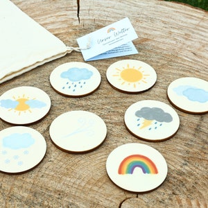 May include: A set of eight wooden weather themed circles with images of a sun, clouds, rain, snow, wind, lightning, and a rainbow. The circles are arranged on a wooden surface with a small white bag in the background.