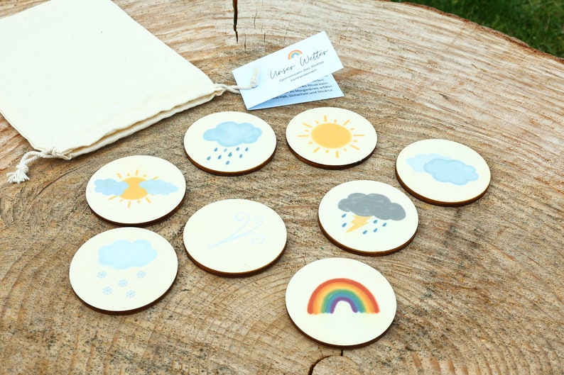 May include: A set of eight wooden weather themed circles with images of a sun, clouds, rain, snow, wind, lightning, and a rainbow. The circles are arranged on a wooden surface with a small white bag in the background.