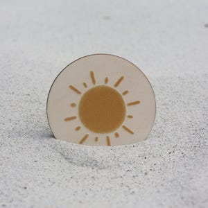 May include: A round, white coaster with a yellow sun design. The sun has a simple, hand-drawn style with rays extending from the center.