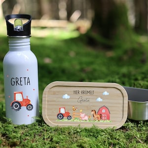 Könnte beinhalten: Eine weiße Wasserflasche und eine Brotdose aus Holz mit einem Edelstahlbehälter. Die Wasserflasche hat einen schwarzen Deckel, den Namen "Greta" und ein rotes Traktormotiv. Die Brotdose hat eine Bauernhofszene.