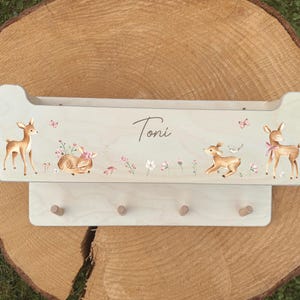 Könnte beinhalten: Hellgrünes Wandregal aus Holz mit einem dekorativen Design mit Rehen, Blumen und Schmetterlingen. Das Regal hat drei Holzstifte darunter zum Aufhängen von Gegenständen. Der Name "Toni" ist auf dem Regal geschrieben.