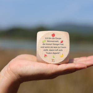 Pode incluir: Um dado de madeira com texto e ilustrações de frutas e vegetais. O texto diz: "Ich bin die Raupe Nimmersatt, die immer Hunger hat, und wenn sie was zu Essen sieht, dann ruft sich 'Guten Appetit'."