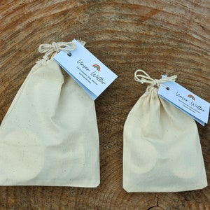 May include: Two white cotton drawstring bags with a small tag that says "Unser Witter" with a rainbow graphic. The larger bag is on the left and the smaller bag is on the right. Both bags are sitting on a wooden surface.