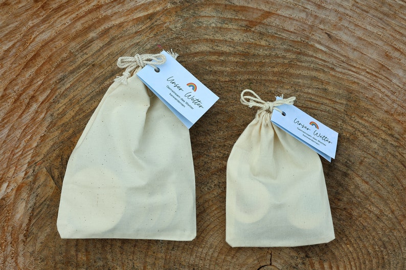 May include: Two white cotton drawstring bags with a small tag that says "Unser Witter" with a rainbow graphic. The larger bag is on the left and the smaller bag is on the right. Both bags are sitting on a wooden surface.
