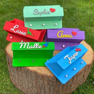 Könnte beinhalten: Fünf bunte Holz-Schlüsselhalter mit Haken, jeder mit einem anderen Namen und einem kleinen Dekoelement. Die Namen sind Sophie, Lasse, Anna, Malte und Jasper. Die Dekoelemente sind ein Herz, ein Dinosaurier, ein Herz, eine Rakete und ein Auto.