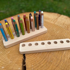 May include: A wooden pencil holder with colorful pencils. The holder is rectangular with holes to hold the pencils upright. A second, empty holder is also visible. The pencils are various colors, including blue, yellow, green, and brown.