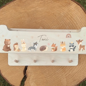 May include: A light-colored wooden wall shelf with a row of illustrated woodland animals and the name "Toni" in script. Below, there are four wooden pegs for hanging items. The shelf is decorated with stars and a rainbow.