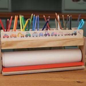 Könnte beinhalten: Holz-Bastelutensilien-Organizer mit einer Rolle weißem Papier und einer Reihe farbiger Bleistifte. Der obere Bereich hat Fächer für Bleistifte und andere Utensilien, verziert mit Fahrzeugillustrationen. Die Basis hat einen roten Akzent.