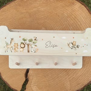 Op de afbeelding: Een lichtgekleurd houten wandrek met een decoratief dierenthema. Het rek heeft een giraf, leeuw, aap en beer, samen met een vliegtuig. De naam "Elisa" staat op het rek geschreven. Inclusief drie houten pinnen om items op te hangen.