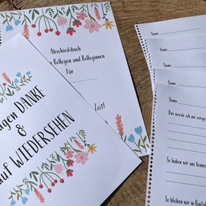 Può includere: Un libro di addio bianco con un bordo floreale e il testo "Wir sagen DANKE & auf WIEDERSEHEN" in nero. Il libro ha una rilegatura a spirale ed è aperto su una pagina con il testo "Abschiedsbuch Kolleginnen und Kollegen für Zeit!" in nero.