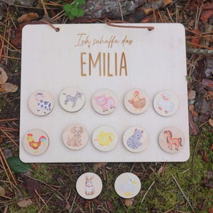 Op de afbeelding: Licht houten bord met de naam "EMILIA" en de zin "Ich schaffe das" in gouden letters. Het heeft een leren koord om op te hangen en 12 houten schijven met illustraties van boerderijdieren, waaronder een koe, een ezel, een varken en een kip.