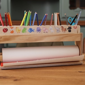 Könnte beinhalten: Holz-Bastelutensilien-Organizer mit einer Papierrolle und einem oberen Bereich für Bleistifte und Buntstifte. Der obere Bereich ist mit bunten Tieraufklebern verziert. Der Organizer ist aus hellem Holz gefertigt.