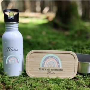 Könnte beinhalten: Eine hellblaue Wasserflasche, eine Brotdose aus Holz und ein Edelstahlbehälter liegen auf Moos. Die Wasserflasche und die Brotdose zeigen ein Regenbogen-Design und den Namen "Marlo". Der Deckel der Brotdose trägt den Text "Sei frech, wild und wunderbar Marlo".