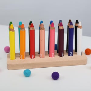 Pode incluir: Um porta-lápis de madeira com uma fileira de lápis coloridos e várias bolinhas de feltro. Os lápis são de várias cores, incluindo amarelo, laranja, vermelho, rosa e azul. As bolinhas de feltro são em tons de rosa, verde, azul, laranja e amarelo.