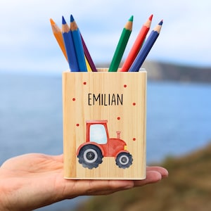 May include: A wooden pencil holder with a red tractor illustration and the name "EMILIAN" printed on the front. The holder contains several colored pencils. The background is a blurred view of the ocean and a hillside.