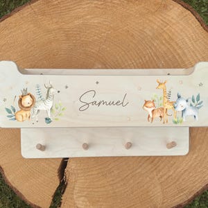 Op de afbeelding: Houten wandplank met een lichtgekleurde afwerking, met een geschilderd safari-dierenontwerp. De plank bevat de naam "Samuel" in script en vier houten pinnen eronder. Het ontwerp omvat een leeuw, zebra, giraffe en andere dieren.