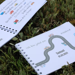 Könnte beinhalten: Ein weißes Heft mit Spiralbindung zeigt eine Straße mit Autos und den Text "Ich bin jetzt ein Kitakind Samuel 01.09.2024".