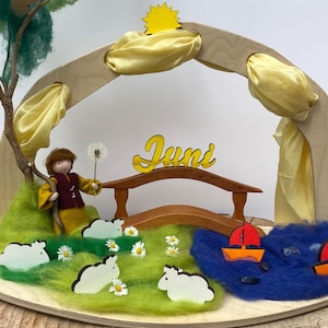 Könnte beinhalten: Eine Holzspielszene mit einer gelben Sonne, einem gelben Stoffbogen, einer Holzbrücke mit dem Wort "Juni" darauf, einer Schäferfigur, weißen Schaffiguren und blauem Wasser mit zwei roten Segelbooten.