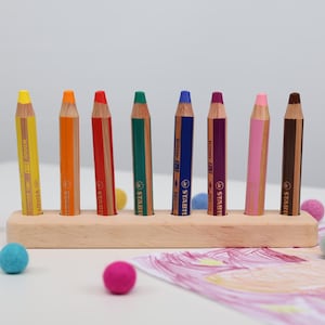 Peut inclure: Un porte-crayons en bois avec huit crayons de couleur. Les crayons sont jaunes, orange, rouges, verts, bleus, violets, roses et marron. Des boules de feutre colorées et un dessin sont éparpillés autour du support.