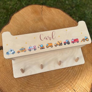 Könnte beinhalten: Holzregal mit dem Namen "Carl" und bunten Fahrzeugillustrationen. Es zeigt einen Polizeiwagen, ein Taxi, einen Krankenwagen, einen Bagger, einen Muldenkipper, einen Betonmischer, einen Traktor, ein Feuerwehrauto und einen Frontlader.