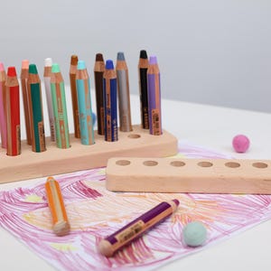 May include: A wooden pencil holder with colorful pencils. The pencils are arranged in a light-colored wooden holder. There are also loose pencils and colorful felt balls scattered around. The pencils are various colors, including yellow, pink, red, and blue.